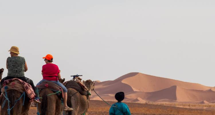 Caravana de camellos con jinetes se dirige hacia las dunas de arena ondulantes del Sahara al atardecer.