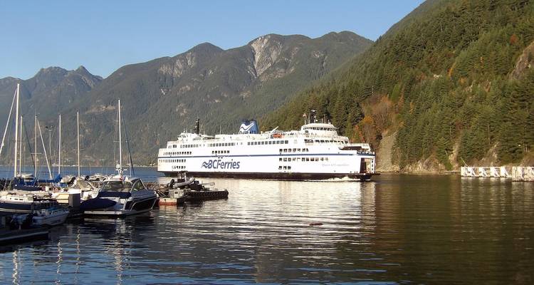 Large BC Ferries vessel passing forested mountains and marina boats on a calm inlet.