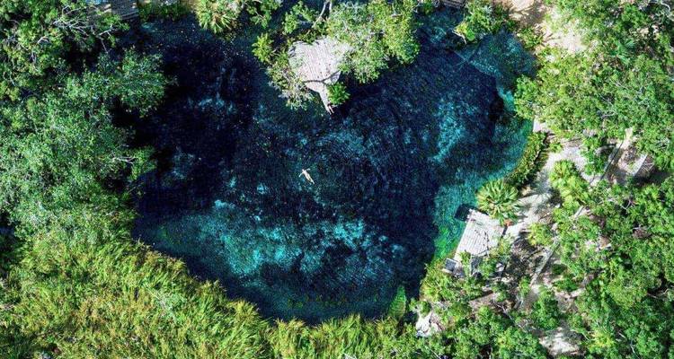 Vue aérienne d'un cénote d'un bleu éclatant entouré d'une jungle dense avec un nageur solitaire au centre.