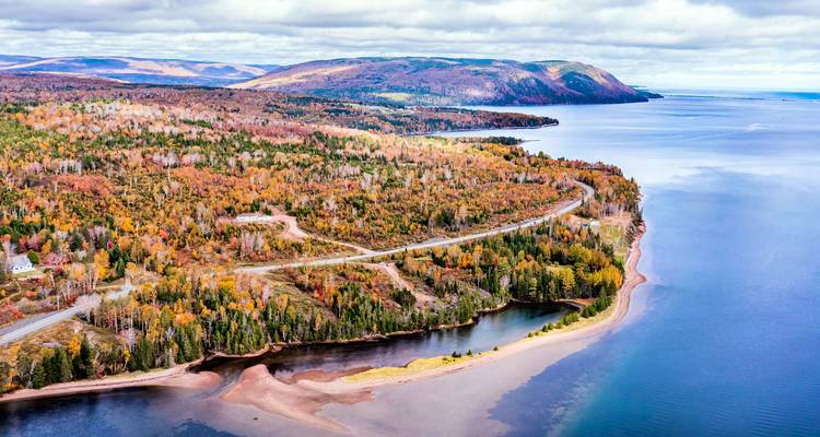 Une vaste forêt d'automne rencontre une route côtière sinueuse bordée d'eaux bleues calmes et de collines lointaines sur l'île du Cap-Breton.