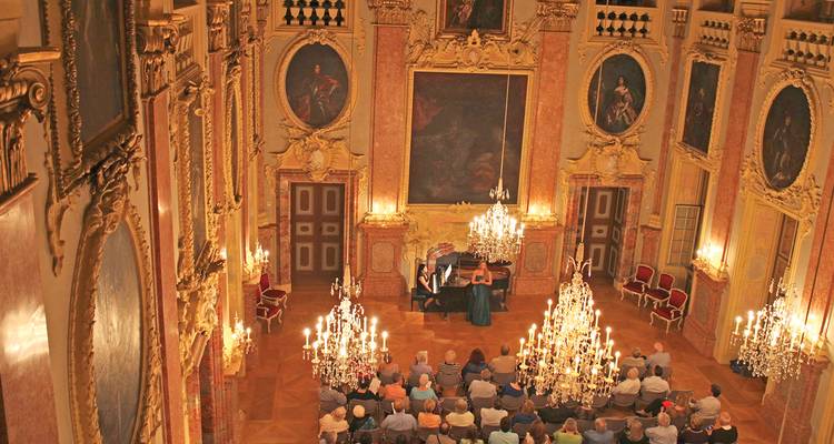 Gasten zittend in een sierlijke paleiszaal die een klassiek muziekoptreden bekijken onder grote kroonluchters