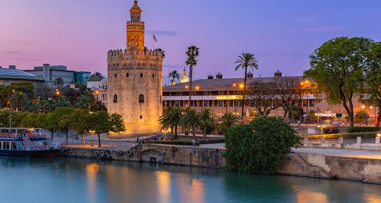 Der beleuchtete Torre del Oro und die Flusspromenade spiegeln sich bei Einbruch der Dämmerung in ruhigem Wasser mit violettem Himmel.