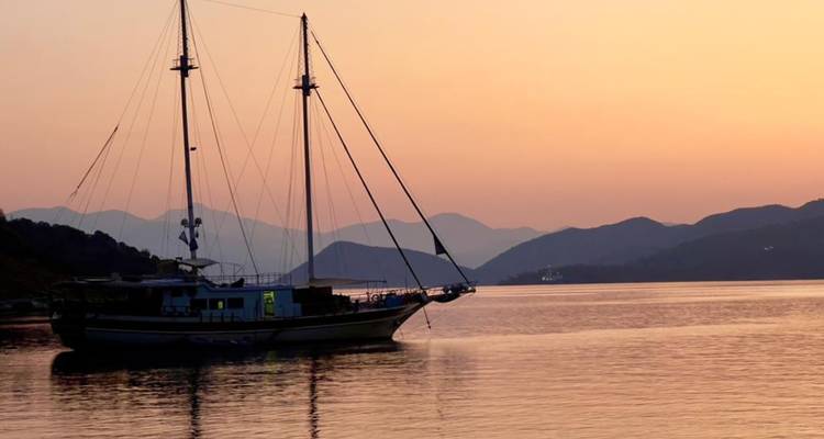 Voilier ancré sur une eau calme avec des silhouettes de montagnes en couches et un ciel du crépuscule orange.