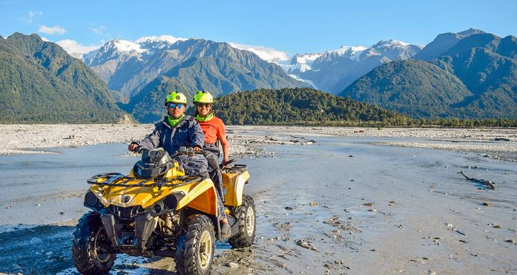 Deux cavaliers sur un véhicule tout-terrain jaune traversent un large lit de rivière avec des montagnes aux sommets enneigés en arrière-plan