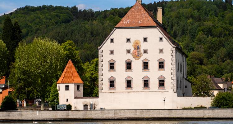 Historisch huis aan de rivier met een geschilderde gevel tegen groene beboste heuvels.