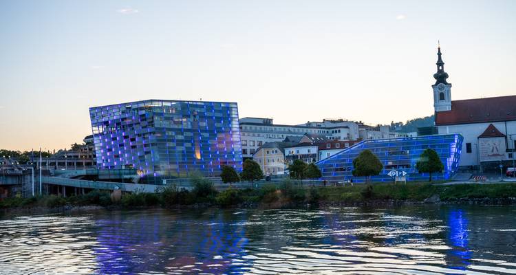 Vue en soirée du Musée d'art Lentos de Linz illuminé en bleu, se reflétant sur le Danube