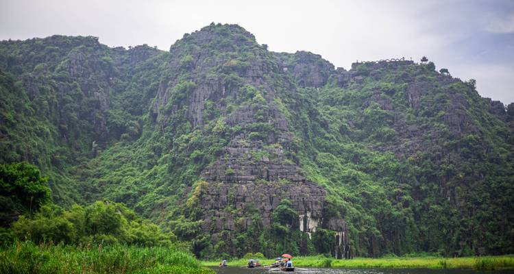 Steep limestone karst cliffs covered in greenery rise above boats on a tranquil river valley