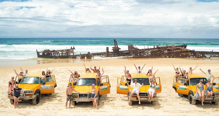 Opgewonden reizigers poseren op gele 4WD's naast het verroeste scheepswrak de Maheno op het strand van Fraser Island.