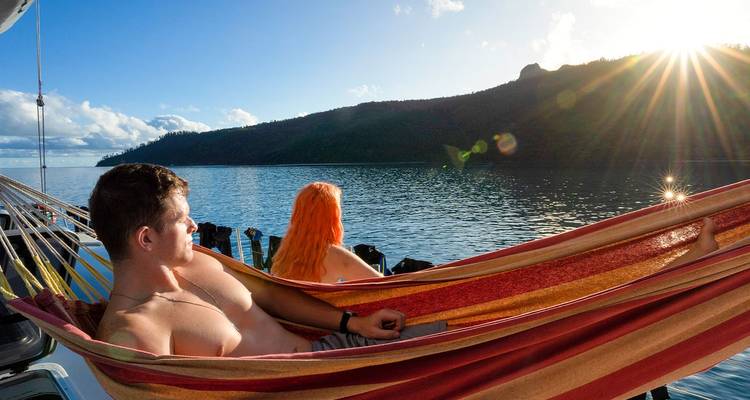 Pareja relajándose en una hamaca colorida en un velero con rayos de sol sobre una bahía tranquila.