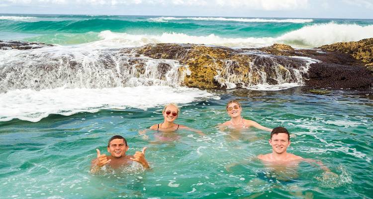 Cuatro amigos disfrutando de una piscina natural de roca mientras las olas se estrellan detrás de ellos.