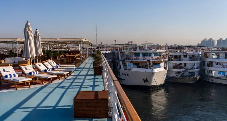 Sun-lit deck with loungers on a river cruise ship, moored beside several white riverboats.
