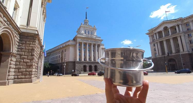 Une main tenant une brillante casserole en métal devant l'imposant bâtiment gouvernemental Largo de Sofia par une journée ensoleillée.