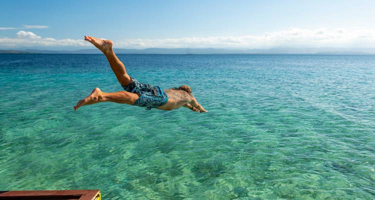Un homme en short de bain plonge avec élégance d'une plateforme en bois dans une mer turquoise cristalline.