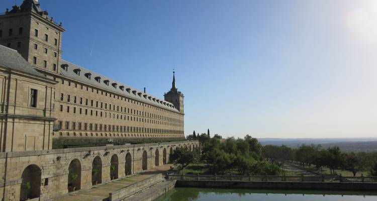 Lange Fassade des Klosters El Escorial mit Arkadenbogen über einem reflektierenden Teich unter hellem Himmel.