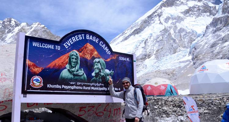 Wanderer zeigt auf das Schild des Everest Base Camp mit dem schneebedeckten Khumbu-Gletscher und Zelten im Hintergrund.