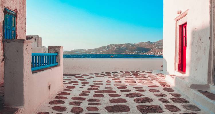 Callejón cicládico con pavimento de piedra, paredes encaladas, balcón azul brillante y vistas al mar a lo lejos.