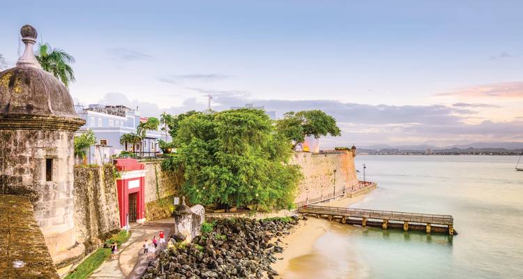 Fuerte histórico junto al mar y edificios coloniales en tonos pastel se asoman a aguas turquesas tranquilas y una pequeña playa de arena al atardecer en San Juan.