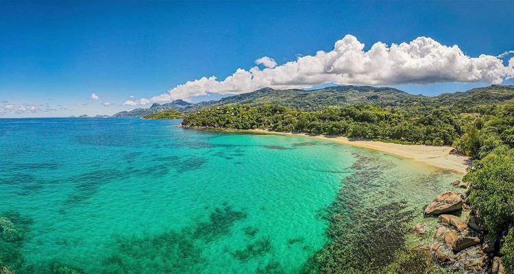 Plage tropicale des Seychelles avec eau turquoise, récifs coralliens et jungle montagneuse en arrière-plan.