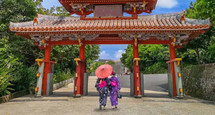 Two women in colorful kimonos with parasol walk through a traditional red gate in Okinawa.