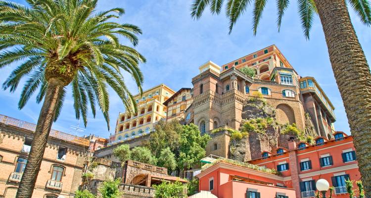 Historic cliff-top buildings framed by tall palm trees against a bright summer sky on Italy’s Amalfi Coast.