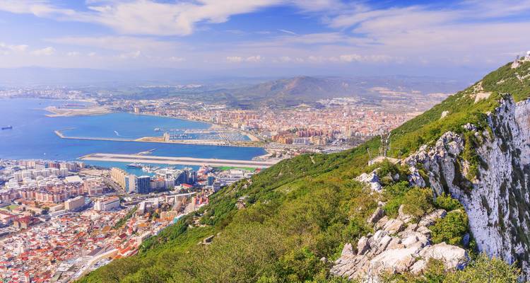 Vista aérea de la Roca de Gibraltar mostrando la ciudad, el puerto y la pista de aterrizaje bajo un cielo azul brillante.