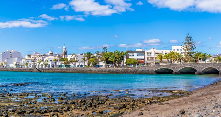 Pueblo costero de Arrecife con edificios blancos y un puente de piedra con arcos reflejado en agua turquesa.