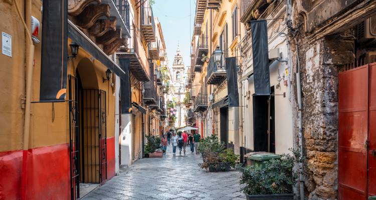 Ruelle étroite et colorée avec balcons et plantes, menant à un clocher d'église au loin tandis que les piétons se promènent sur les pavés.