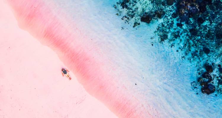 Vista con dron de una orilla de arena rosa vibrante que se desvanece en agua clara con rayas de arrecife y un solitario tomador de sol.