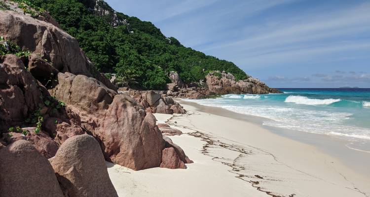 Plage de sable blanc isolée bordée de rochers de granit rose et de verdure de jungle avec des vagues douces.
