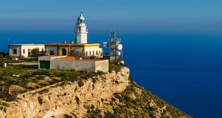 Faro blanco costero encaramado en lo alto de un acantilado escarpado con vistas al profundo mar Mediterráneo azul.