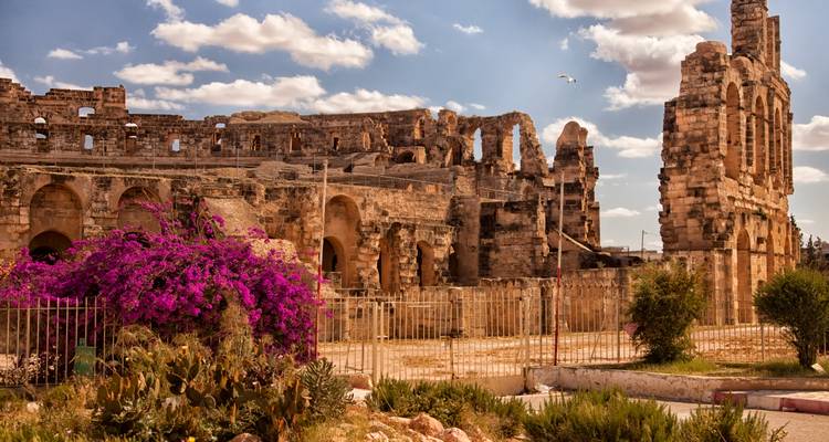 Ruinas de un anfiteatro romano antiguo enmarcadas por bugambileas vibrantes bajo un cielo parcialmente nublado.