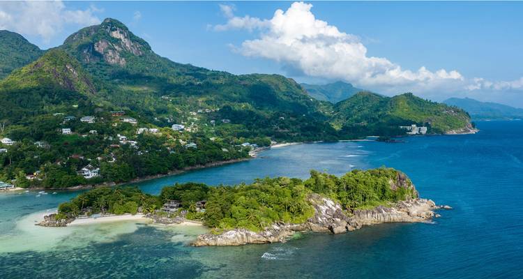 Vue aérienne du littoral luxuriant et verdoyant de Mahé avec un minuscule îlot boisé et des baies turquoise sous un ciel bleu éclatant.