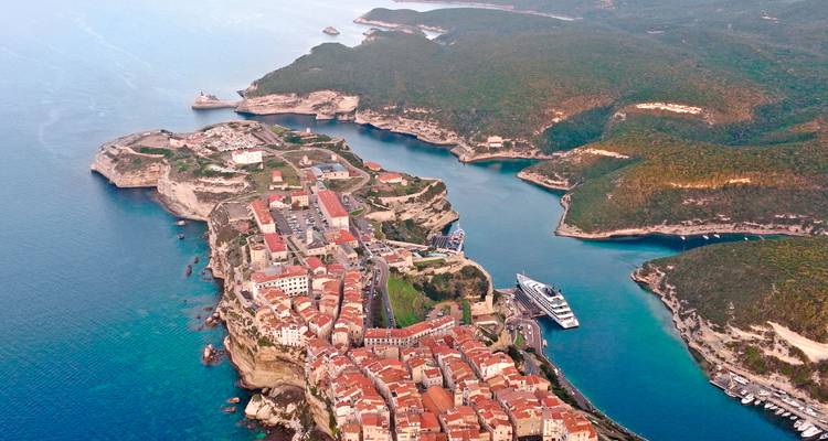 Vue aérienne spectaculaire de la ville de Bonifacio perchée sur les falaises avec ses toits en terre cuite et un yacht pénétrant dans le port étroit aux eaux turquoise en forme de fjord.