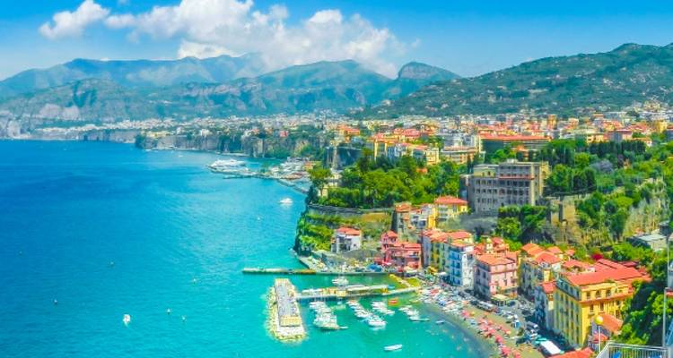 La ville côtière colorée s'étend le long de la baie de Naples d'un bleu éclatant, encadrée par des montagnes vertes et escarpées sous des nuages épars.