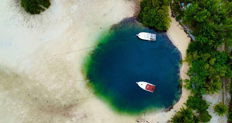 Aerial view of a deep blue cenote-like lagoon connected to a sandy tidal flat with two small boats moored.