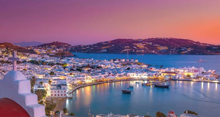 Vista al atardecer sobre el puerto de Mykonos con casas encaladas, molinos de viento y barcos brillando bajo cielos púrpuras.