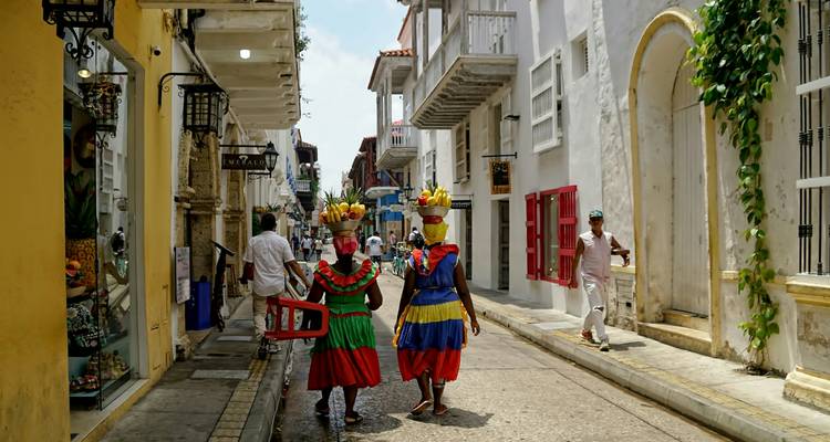 Des palenqueras traditionnelles vêtues de robes colorées marchent le long d'une rue historique bordée de bâtiments aux couleurs pastel.