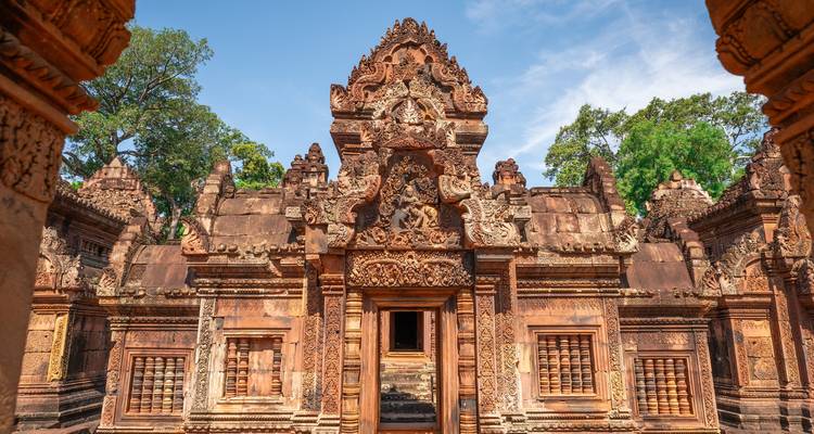 Façade de temple en grès rouge finement sculptée, entourée d'arbres luxuriants sous un ciel bleu.