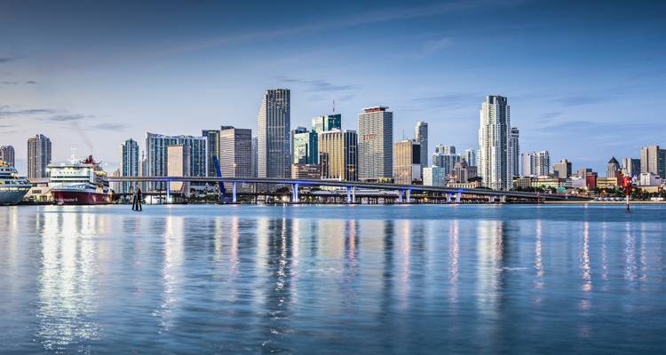 Les eaux réfléchissantes de la baie de Biscayne reflètent l'horizon urbain du centre-ville de Miami sous un ciel bleu du début de soirée.