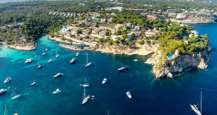 Vista aérea de una cala turquesa salpicada de yates a vela y una punta cubierta de pinos en Mallorca.