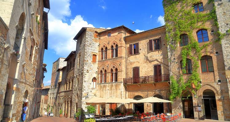 Charming stone street lined with ivy-clad buildings and an inviting outdoor café beneath blue summer skies.