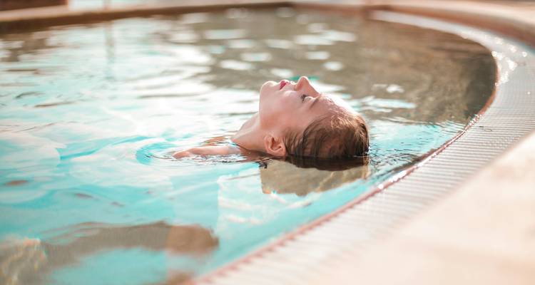 Woman floats serenely on her back in a warm outdoor pool, head tilted to sky.