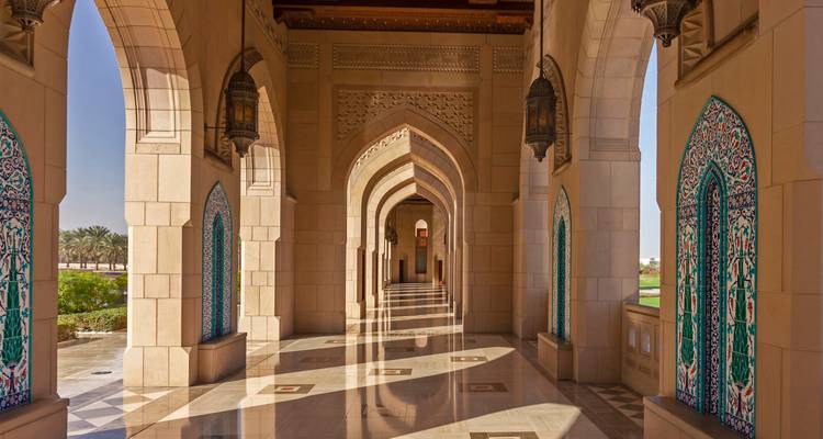 Marmorkorridor der Sultan-Qabus-Moschee mit verzierten Bögen und Mosaikpaneelen, die sich auf dem polierten Boden spiegeln.