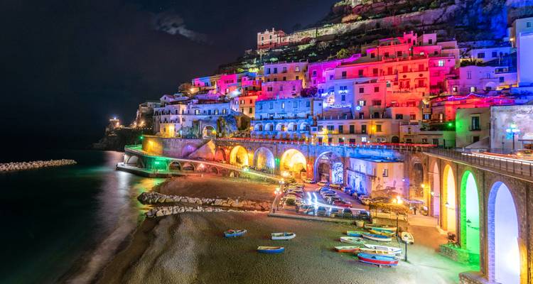 Le village perché sur les falaises s'illumine de lumières multicolores se reflétant sur une plage tranquille et la mer la nuit.
