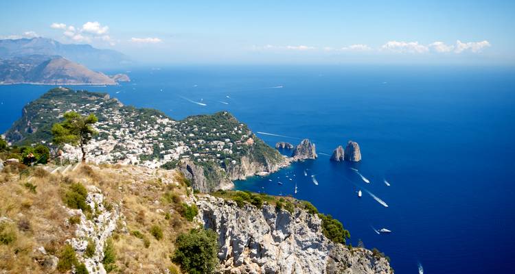 Panorama dramatique en haut de la falaise surplombant l'île de Capri, les formations rocheuses marines de Faraglioni et la mer Méditerranée bleu profond parsemée de bateaux.