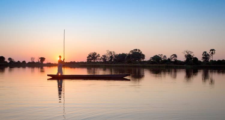 Silhouetted guide standing on a mokoro canoe at sunrise on a calm lagoon
