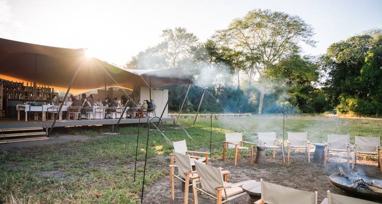 Freiluftkampf-Speisebereich mit Morgenlicht und Stühlen um eine rauchende Feuerstelle.