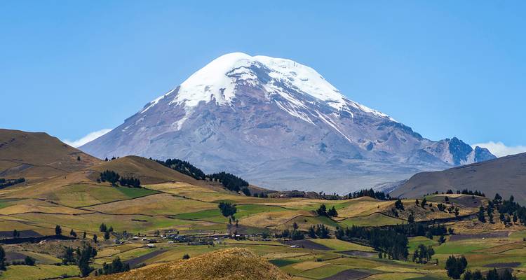 Der schneebedeckte Vulkan Chimborazo erhebt sich über ein buntes Flickenteppich-Farmland unter klarem blauen Himmel.