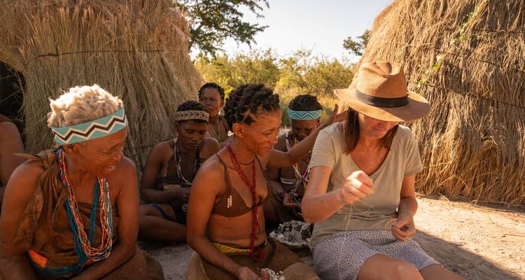 Besucher lernt traditionelles Handwerk von indigenen San-Frauen, die neben strogedeckten Hütten sitzen.
