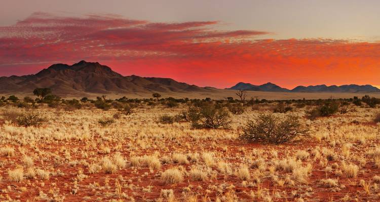 Weitläufige Kalahari-Landschaft mit roter Erde und dramatischem Sonnenuntergangshimmel über entfernten Hügeln.
